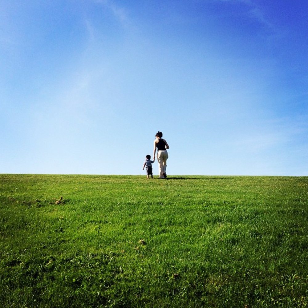 A mother and her young son walking hand in hand up a grassy hill under a blue sky